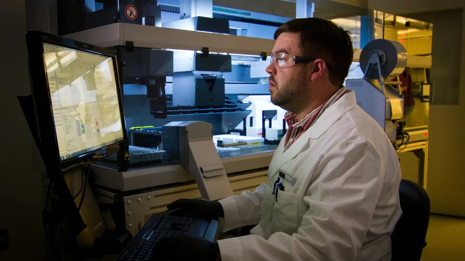 Laboratory technician in a white coat and safety glasses reviewing data on a monitor next to an automated laboratory system