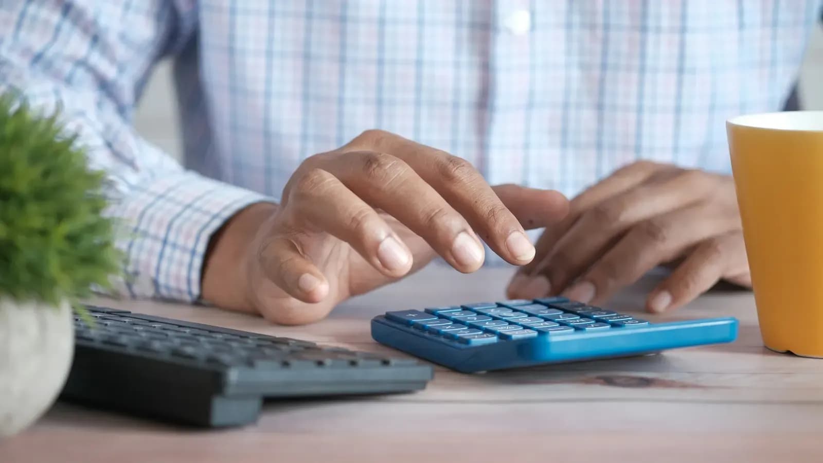 Close-up of a person's hands using a blue calculator on a wooden desk next to a keyboard and a yellow mug