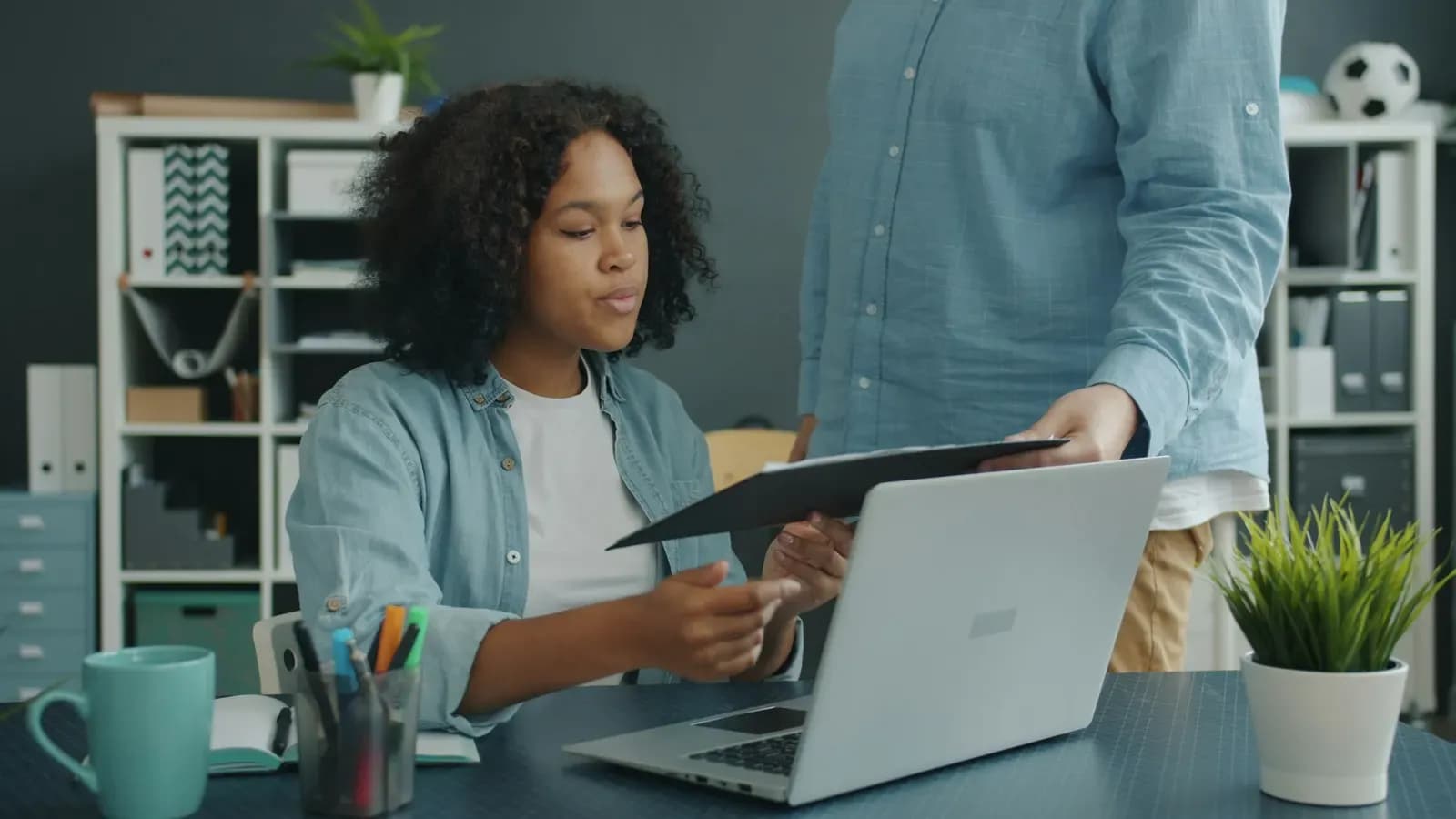 Two colleagues at an office desk, one handing the other a document folder next to a laptop