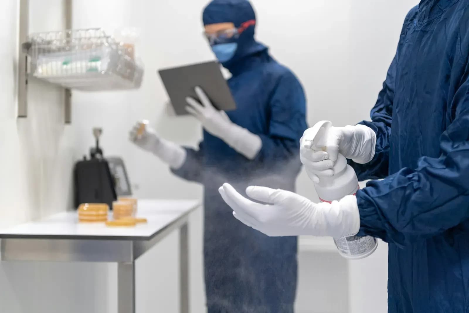 Two cleanroom technicians in blue gowns: one sprays disinfectant on gloved hands while the other reads a tablet