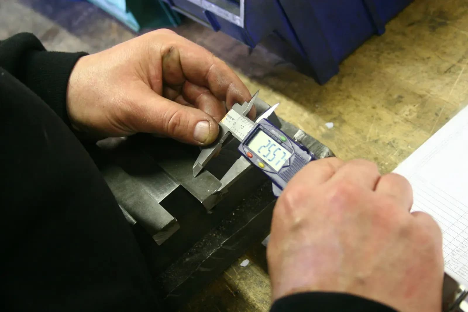 Hands measuring a machined metal part with a digital caliper next to an inspection record sheet